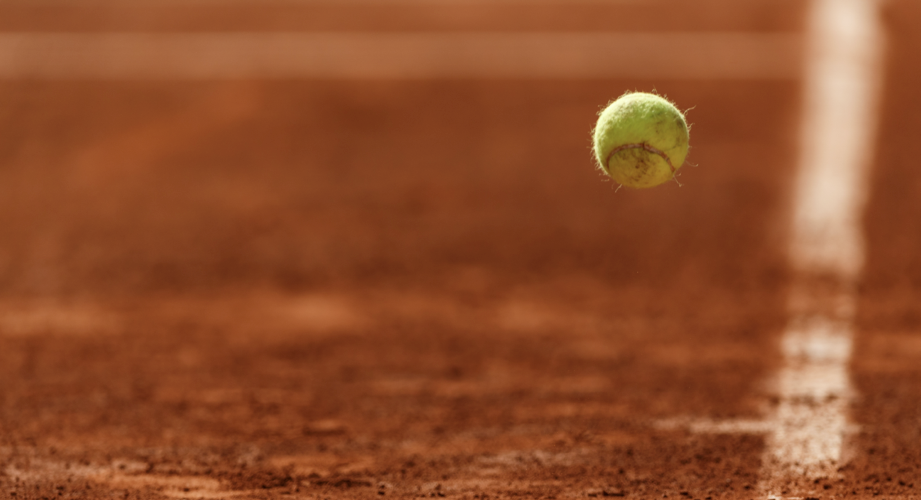 A close up of a tennis ball suspended in mid air against a red clay pitch