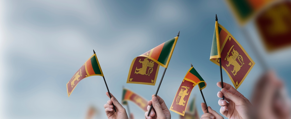 Hands holding small Sri Lankan flags aloft against a blue sky