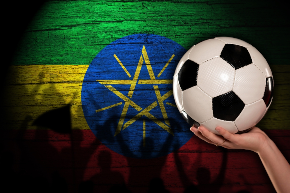 Hand holding a football in front of the Ethiopian flag painted on wooden boards, with silhouettes of fans below