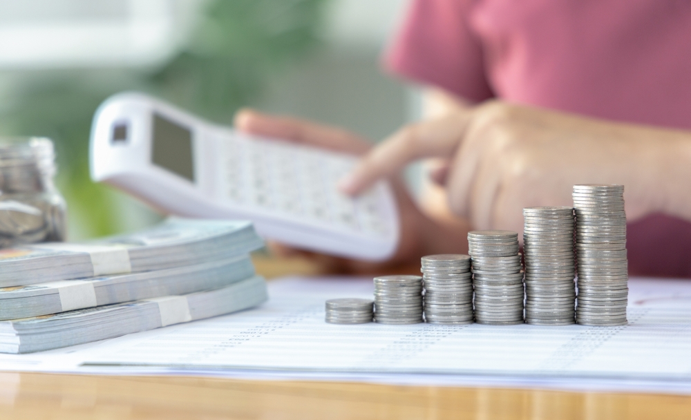 Counting coins and banknotes with a calculator Ascending stacks of coins on a printed spreadsheet, with a person using a calculator and bundles of banknotes in the background