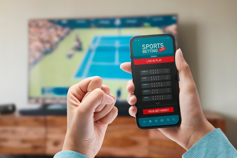 Live in-play tennis betting on a smartphone Hands holding a smartphone showing a live in-play tennis betting screen, with a match on TV behind