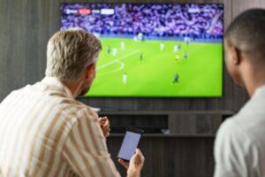 Two people watching a football match on television while holding a smartphone
