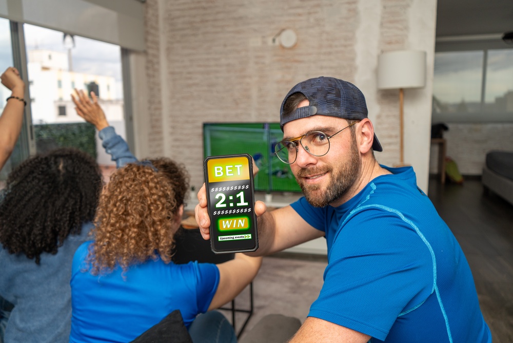 Live betting on a smartphone while watching football with friends Man holding up a smartphone displaying a betting app while friends watch football on TV